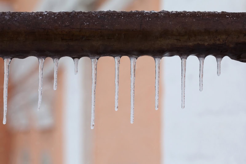 What Should I Do to Fix Frozen Pipes? Icicles hanging from a brown pipe. Frozen water and metal surface, winter time concept. selective focus shallow depth of field photo.